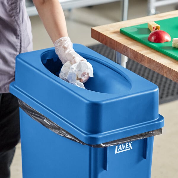 A woman in gloves putting a plastic bag in a dark blue Lavex slim rectangular trash can.