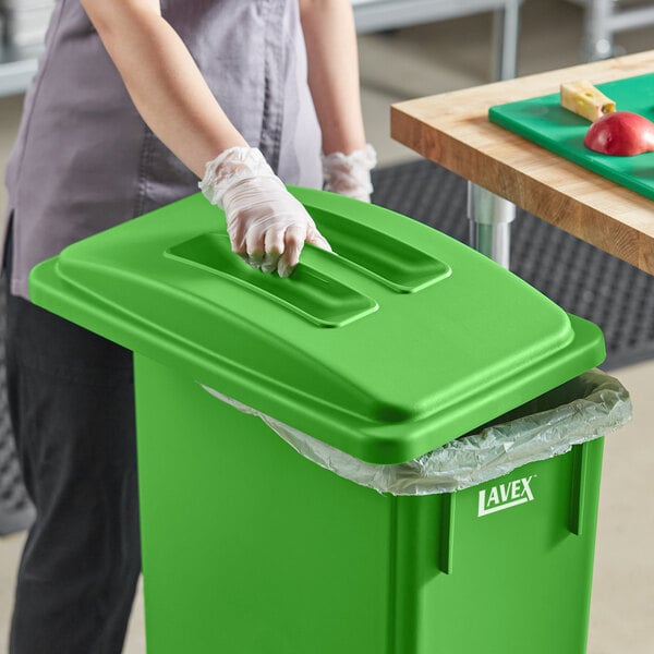 A woman in an apron and gloves putting a Lavex Lime Green Slim Rectangular Compost Receptacle with Flat Lid into a kitchen counter.