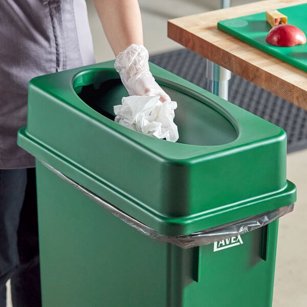 A woman in gloves using a Lavex dark green slim rectangular trash can on a counter in a school kitchen.