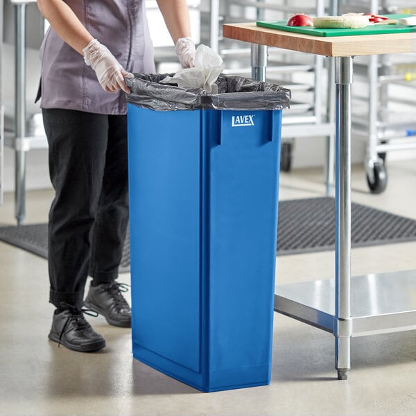 A woman in a blue uniform putting a black garbage bag in a Lavex dark blue rectangular trash can.
