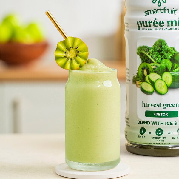 A bottle of Smartfruit Harvest Greens puree next to a glass of green smoothie with a straw.