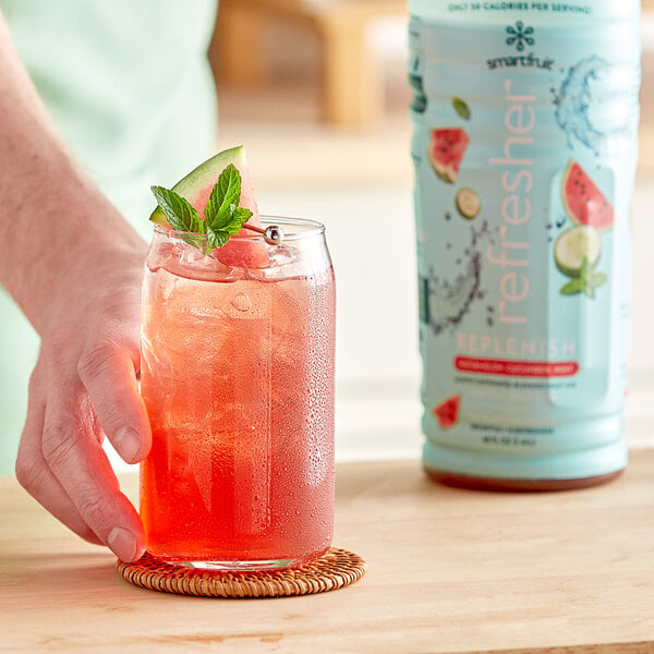 A hand holding a glass of Smartfruit Replenish Watermelon / Cucumber / Mint Refresher Beverage with a mint leaf.