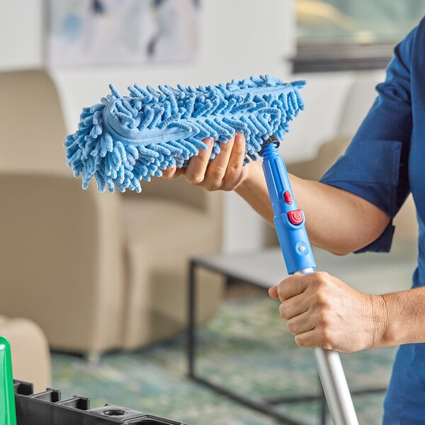 A person is attaching a blue chenille duster head to a blue telescopic pole in a room with beige furniture.