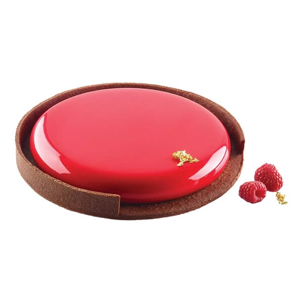 A round silicone baking mold with a glossy red tart and a chocolate crust, accompanied by raspberries and a gold garnish.