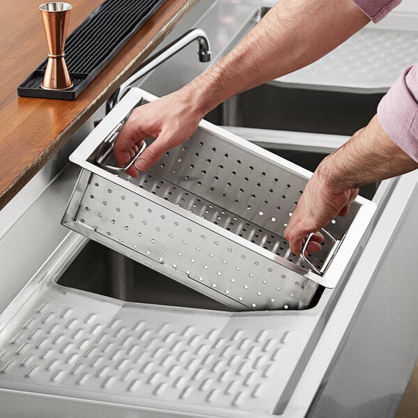 A person using a Regency stainless steel perforated scrap/pre-rinse basket in a sink.