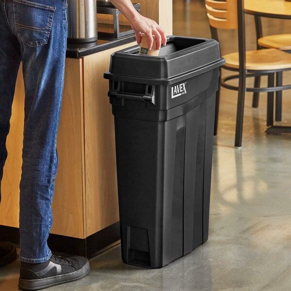 A man leaning against a Lavex Pro black slim rectangular trash can with a black lid.