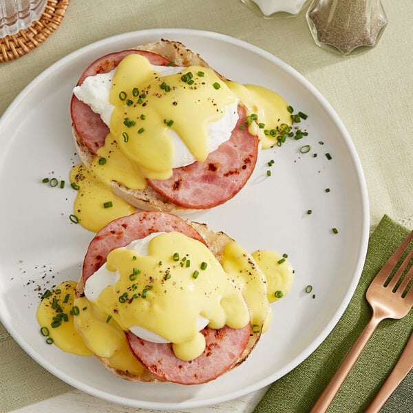 A plate of eggs benedict with ham and cheese on a white plate with a fork and knife.