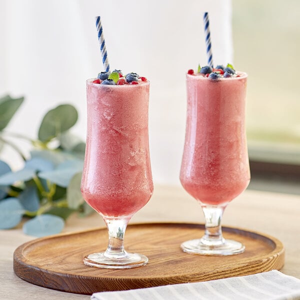 A wooden tray with two glasses of Capora Blueberry Pomegranate smoothies with blue and white straws.