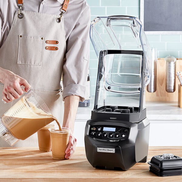 A man pouring a brown liquid into a black Hamilton Beach Summit Edge blender on a counter.