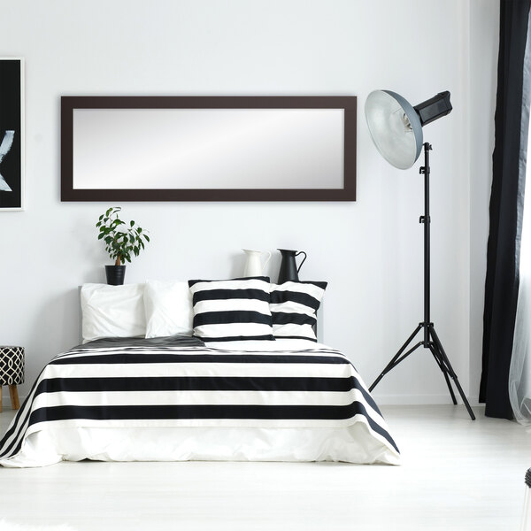 A BrandtWorks matte black mirror leaning against a wall in a white bedroom with a black and white striped bed.