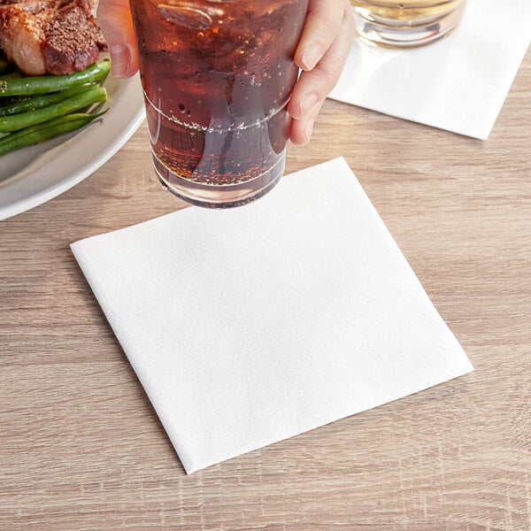 A hand holding a glass of brown liquid over a Touchstone by Choice white beverage napkin on a table.