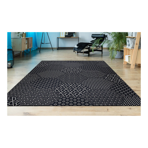 A Couristan Afuera Anode area rug with black and white geometric patterns on a floor in a room with a black chair and a plant.
