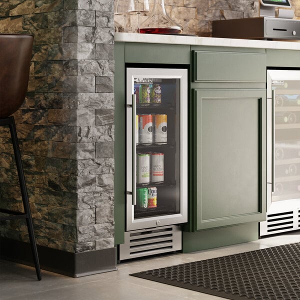 A slim glass door beverage cooler built into green cabinetry, stocked with assorted cans.