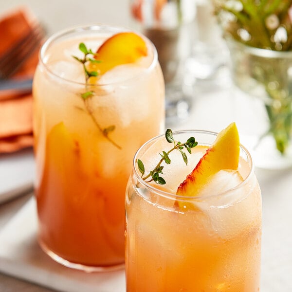 Two glasses of peach juice with ice and fruit slices, garnished with herbs and a slice of peach.