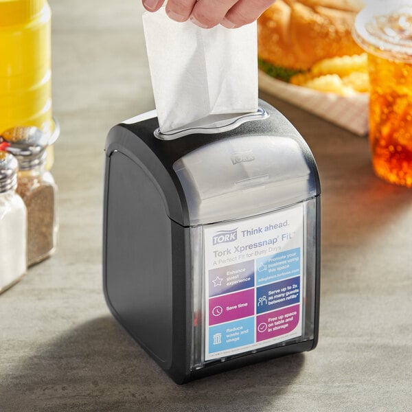 A person using a black Tork Xpressnap tabletop napkin dispenser on a counter.