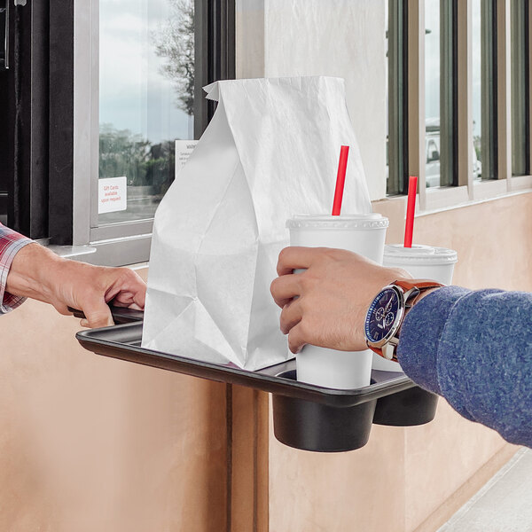 A hand holding an Elite Global Solutions black bamboo and melamine contactless serving tray with drinks and a white bag.