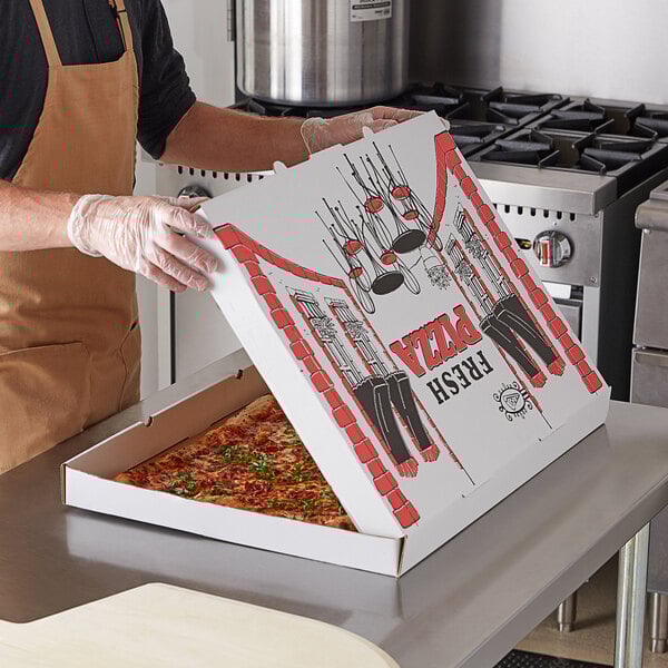 A man in an apron holding a white corrugated pizza box with a pizza in it.