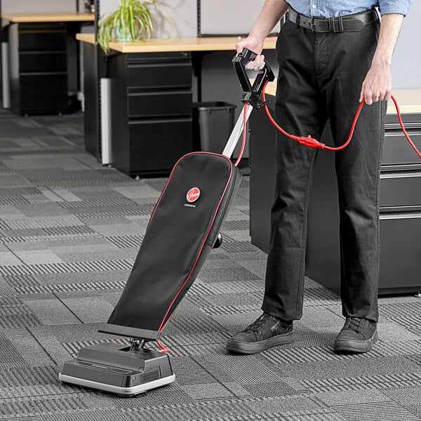 A person holding a Hoover Superior Lite upright vacuum cleaner with a red cable vacuuming a carpet in a corporate office cafeteria.