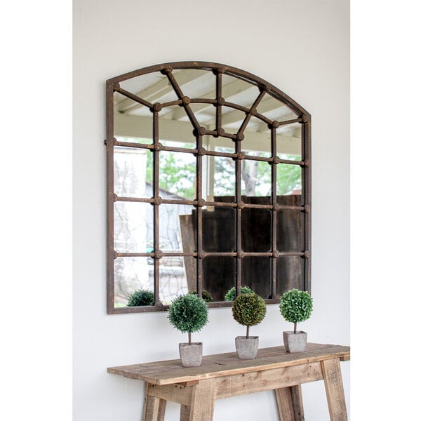 A Kalalou arched iron mirror on a wall over a wooden bench with two small round trees in pots.