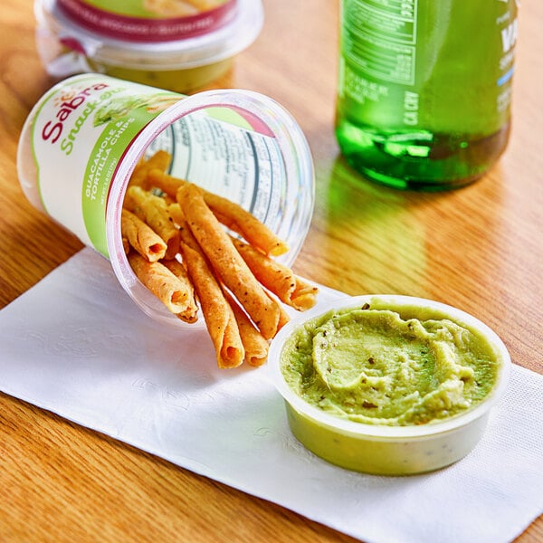 A small container of Sabra guacamole with rolled tortilla chips on a table.