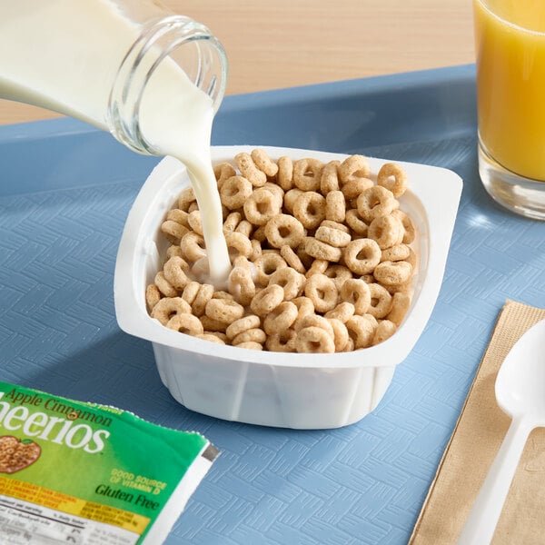 A single-serve bowl of Apple Cinnamon Cheerios cereal with milk being poured in, placed on a blue tray next to a glass of orange juice and a packaged cereal label.