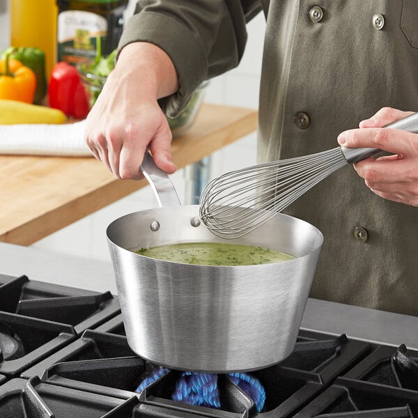 A person stirring green liquid in a Vollrath stainless steel sauce pan on a stove.