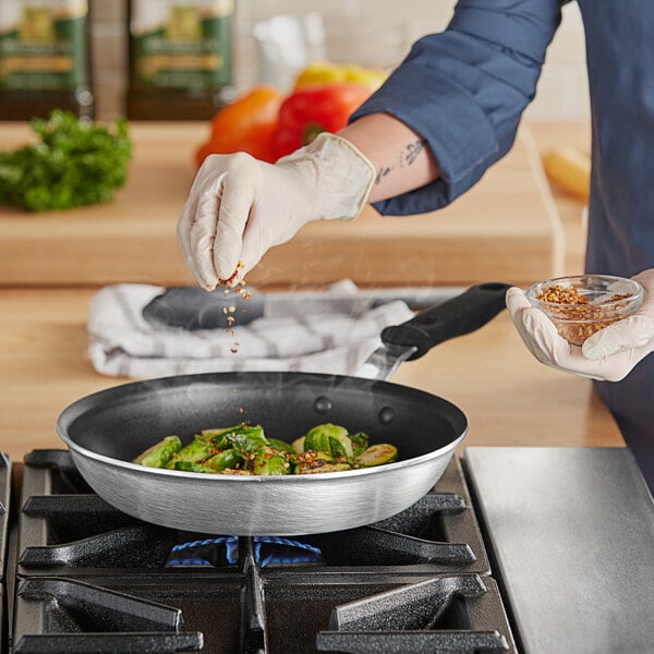 A person in gloves pouring seasoning on food in a Vollrath Wear-Ever non-stick fry pan.