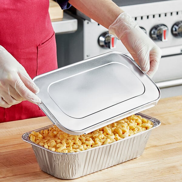 A person in gloves holding a Western Plastics foil steam table tray with macaroni and cheese with the lid open.