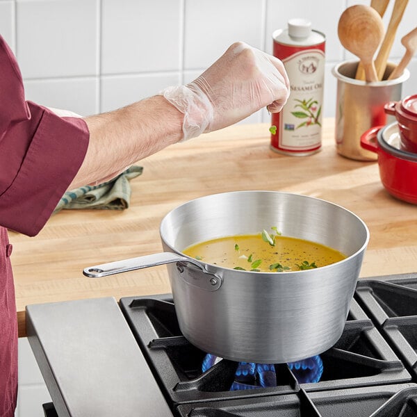 A person cooking soup in a Vollrath stainless steel sauce pan on a stove.