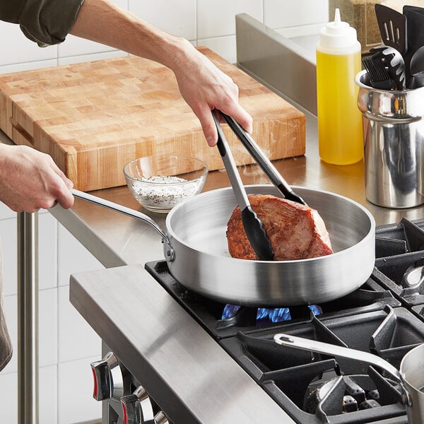 A person using tongs to cook meat in a Vollrath Wear-Ever saute pan on a stove.