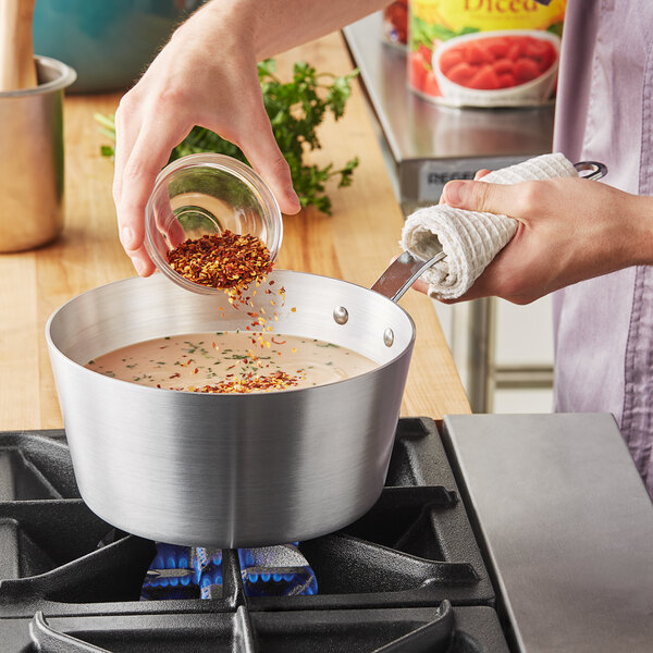 A person pouring sauce into a Vollrath Wear-Ever sauce pan on a stove.