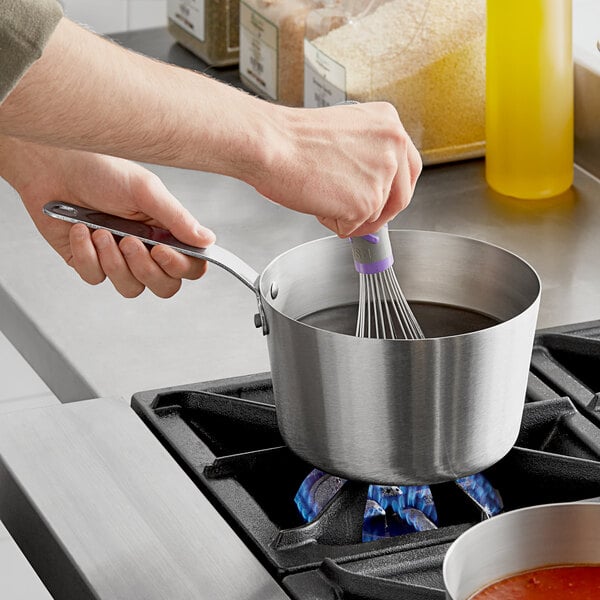 A person stirring yellow liquid in a Vollrath stainless steel saucepan on a stove.