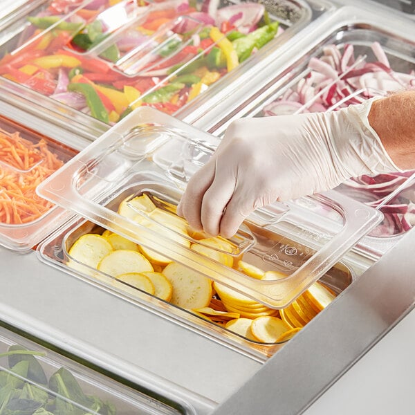 A gloved hand using a clear plastic lid with a handle to cover a food container.