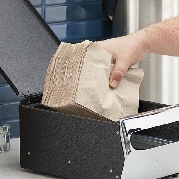 A person's hand putting a brown Dixie paper napkin into a black dispenser.