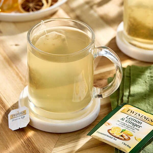 A glass mug of Twinings Lemon & Ginger tea with a tea bag next to it.