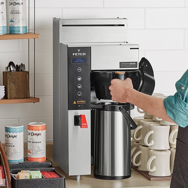 A person using a Fetco coffee machine to pour coffee into a white mug.