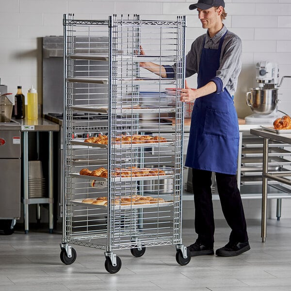 A man in a blue apron pushing a large metal Regency rack of food.