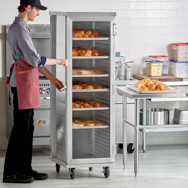 A man wearing an apron standing next to a Regency enclosed sheet pan rack.