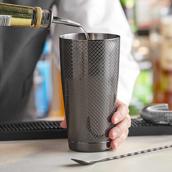 A person using a Barfly black metal cocktail shaker to pour a drink into a cup.