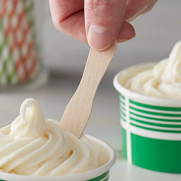 A hand holding a Choice eco-friendly unwrapped wooden taster spoon over a green and white cup of frozen yogurt.