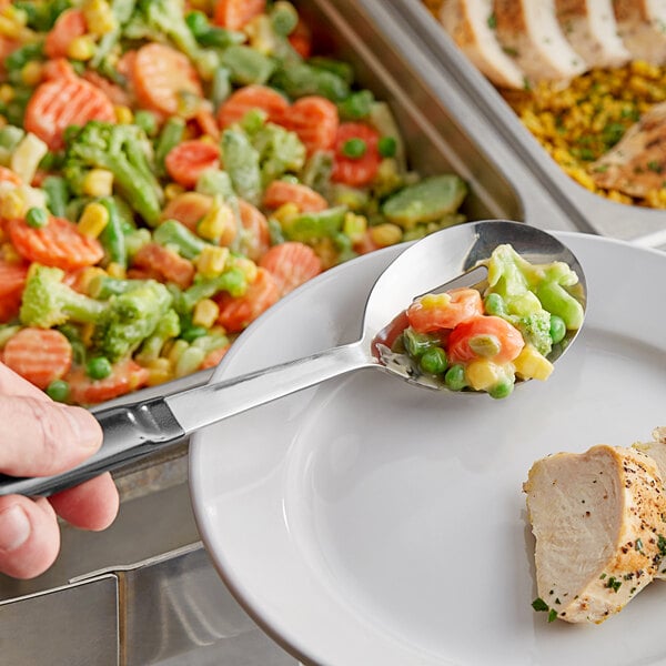 A person holding a Vollrath slotted basting spoon with a black Kool-Touch handle over a plate of vegetables.