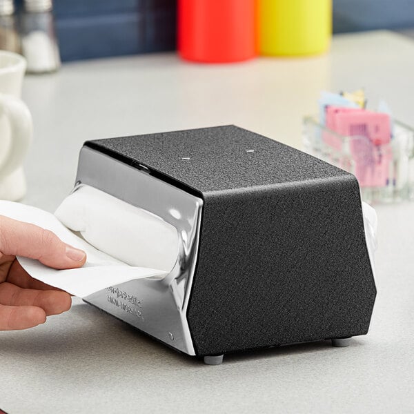 A person using a black Dixie tabletop napkin dispenser with a chrome faceplate.