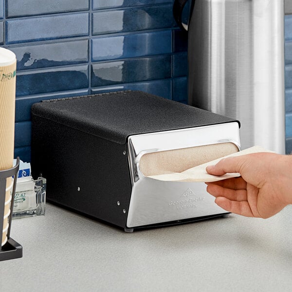 A person using a Dixie black and silver countertop napkin dispenser to get a napkin.