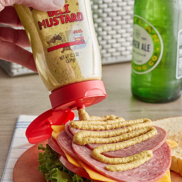 A hand using Mike's Amazing Stoneground Deli Mustard to put mustard on a sandwich.