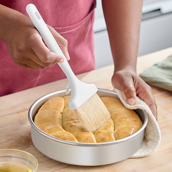 A white-handled boar bristle pastry and basting brush being used to apply glaze to baked bread in a round pan.