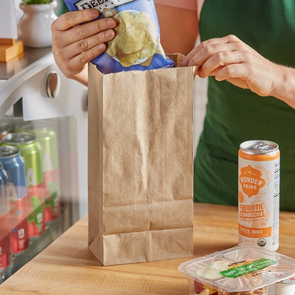 A woman putting a bag of chips into a brown Choice natural kraft paper bag.