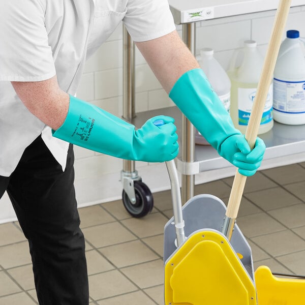 A person wearing green Ansell AlphaTec Solvex gloves and using a yellow mop to clean a floor.