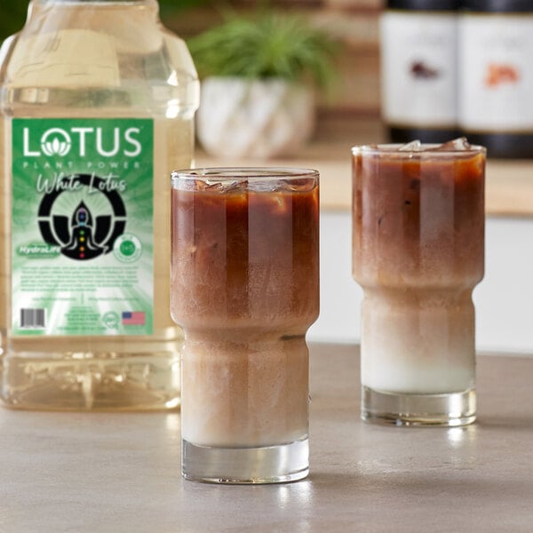 A 64 fl. oz. bottle of Lotus Plant Power White Lotus 5:1 Energy Concentrate is shown next to two iced layered beverages in clear glasses.