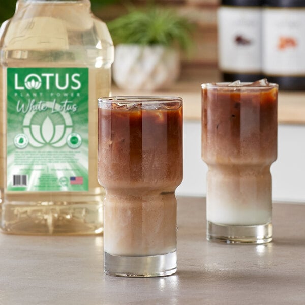 Two iced layered drinks in clear glasses are placed on a countertop, with a large bottle labeled 'Lotus Plant Power White Lotus' in the background.