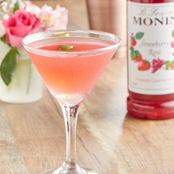 A martini glass filled with pink Monin strawberry rose drink next to a bottle of Monin syrup.